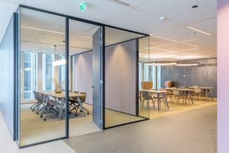 Moderne vergaderruimte met glazen wanden, houten vergadertafel, grijze kantoorstoelen en ronde vaas. Aangrenzend open kantoor