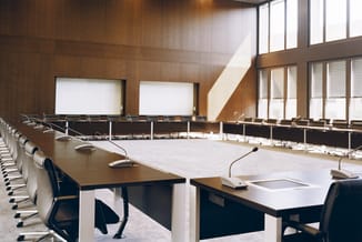 Moderne vergaderruimte met lange houten vergadertafels, zwarte stoelen, microfoons en schermen aan de wand, houten afwerking.