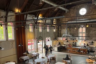 Industrieel interieur met open keuken, houten stoelen, witte tafels, hangende verlichting en grote ramen met rode kozijnen.