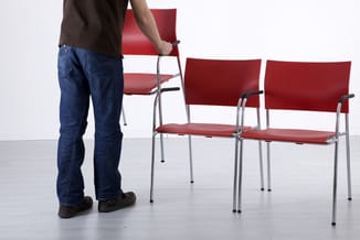 Man plaatst rode stoel met metalen frame en zwarte armsteunen in rij met drie identieke stoelen op betonvloer.