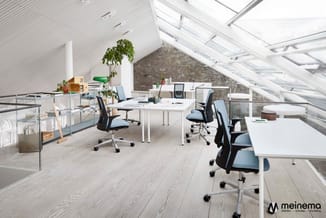 Modern kantoor op zolder met witte bureaus, blauwe ergonomische stoelen, houten vloer, bakstenen muur en grote dakramen.