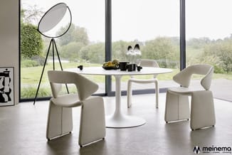 Moderne eetkamer met wit rond tafelblad, drie beige gestoffeerde stoelen, zwarte tripod lamp en uitzicht op groen landschap.