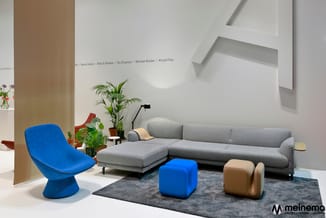 Moderne showroom met grijze hoekbank, blauwe kuipstoel, blauwe en beige poefs op donker tapijt, kolom, plant en wandplanten.