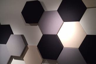 Hexagonale akoestische panelen in zwart, grijs en wit aan muur bevestigd, één paneel verlicht.