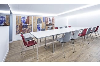 Moderne vergaderruimte met lange witte tafel, rode en grijze stapelstoelen, houten vloer en kunstwerk aan de wand.