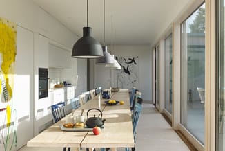 Moderne eetruimte met lange houten tafel, blauwe stoelen, witte keuken, zwarte hanglamp en grote raampartijen.
