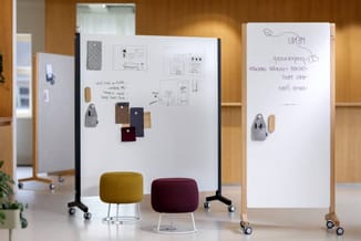 Mobiele whiteboards met schetsen en notities, gele en bordeauxrode poefzitjes met metalen poten, houten kantooromgeving.