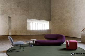 Moderne showroom met paarse gebogen bank, grijze stoel, rode poef en kleine tafels op groen tapijt onder betonnen wanden.