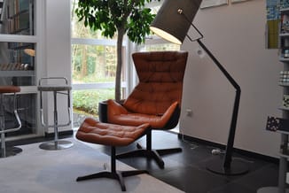 Cognac lederen fauteuil met voetenbankje en zwarte staande leeslamp in moderne kantoorruimte met boekenkasten en raam.