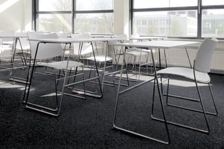 Vergaderruimte met witte kunststof stapelstoelen op grijs tapijt, zwart metalen frames, enkele tafels, grote ramen met uitzic