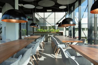 Moderne kantineruimte met houten vergadertafels, grijze stoelen, zwarte hanglampjes met koperen binnenkant en grote ramen met