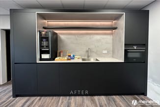 Moderne kantoorkeukeneenheid met zwart front, betonlook achterwand, ingebouwde espressomachine, oven en spoelbak. Tekst "AFTE