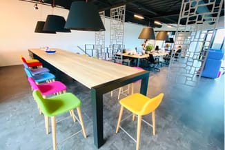Moderne kantoorkantine met zwarte tafel, gekleurde stoelen (groen, geel, roze, blauw, rood), hangende lampen en witte scheidi