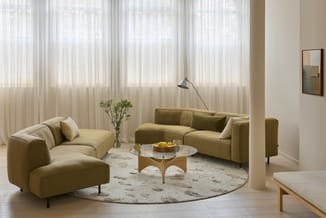 Moderne loungehoek met twee geel-groene fluwelen banken, glazen salontafel op houten onderstel, rond beige vloerkleed, staand
