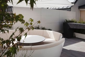 Moderne daktuin met witte ronde zitbank, zwarte tafel, houten vloer, beige zonwering en grijze beplantingsbakken tegen witte