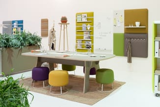 Kantoorruimte met grijze ovale tafel, gekleurde poefzitjes (paars, geel, groen), wandkasten in geel en groen, houten wandelem
