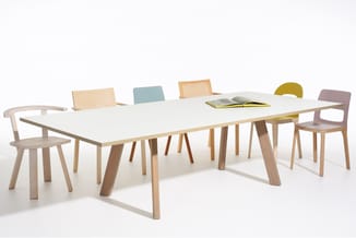 Houten vergadertafel met wit blad en houten onderstel, omgeven door stoelen in verschillende kleuren (hout, blauw, geel, roze