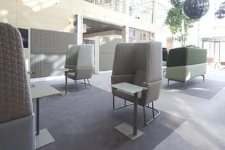 Moderne kantoorruimte met grijze geluiddempende fauteuils met hoge rug aan witte tafels, plant en daglicht.