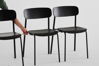 Drie zwarte stoelen met houten rugleuning en zwart zitoppervlak, staande op zwarte metalen poten. Hand plaatst voorwerp op zi