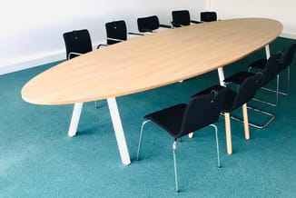 Ovale vergadertafel in licht hout met witte poten, omgeven door zwarte kantooorstoelen in groene vergaderruimte.
