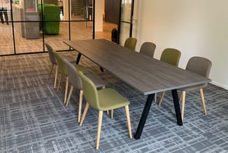 Moderne vergadertafel met donker houten blad en zwart metalen poten, omgeven door stoelen in groen en grijs, in kantoorruimte