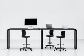 Moderne werkplek met zwart bureau, drie zwarte bureaukstoelen op wielen, monitor, laptop en documenten onder witte wand.