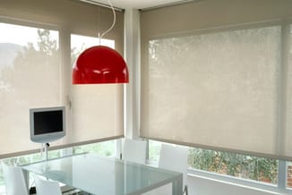 Modern kantoor met glazen tafel, rode hanglamp, beige rolgordijnen en wandmonitor bij raam met uitzicht.