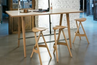 Houten bartafel met witte werkblad en drie houten barkrukken in een industriële kantoorsetting met witte bakstenen muur.