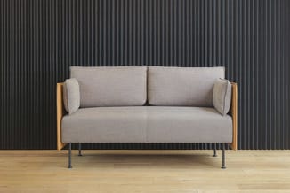 Grijze designbank met houten zijpanelen en metalen poten op houten vloer tegen golfplaten achterwand.