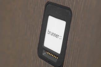 Brunner belcel-display met zwart frame op houten achtergrond, scherm toont 'brunner' logo met contactpunten onderaan zichtbaa