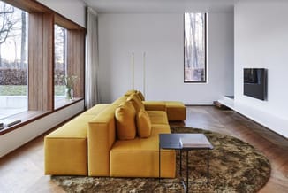 Gele modulaire zitbank met kussens in minimalistisch interieur met houten ramen, donker parket en grijze muren.