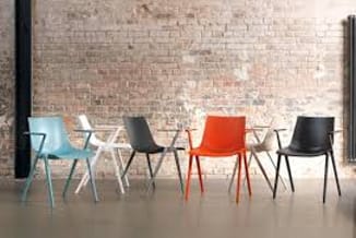 Zes stoelen in verschillende kleuren (blauw, grijs, oranje, zwart) met houten poten tegen bakstenen muur, op betonvloer.