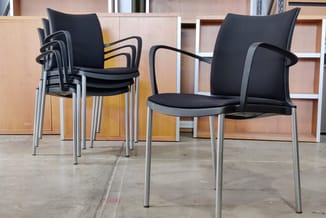 Gestapelde zwarte kantoorstoelen met grijze armleuningen en metalen poten, één stoel vooraan, in een showroom met houten kast