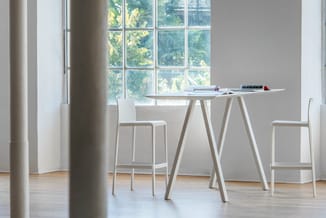 Witte houten werktafel met A-vormig onderstel en twee witte barkrukken in licht kantoor met groot raam.