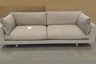 Grijsbeige linnen sofa met twee zitplaatsen, zwevend ontwerp op witte poten, twee armkussens, in tentoonstellingsruimte.