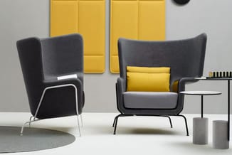Twee grijze loungestoelen met hoge rugleuning in een kantoorruimte. Linkermodel met wit metalen frame, rechtermodel met geel
