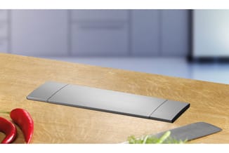 Ergonomische toetsenbord met grijs metallic design op houten bureau, met rode paprika en plant in onscherpe voorgrond.
