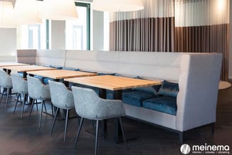 Grijze zitbank met blauwe kussens aan houten tafels, lichtgrijze stoelen, hangende lampen, bruine gordijnen in modern kantoor