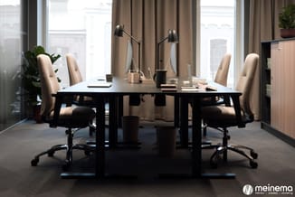 Modern kantoor met zwarte vergadertafel, vier beige bureaustoe­len, zwarte bureaulam­pen en groot raamfront met uitzicht op s