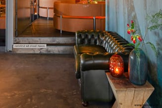 Zwarte Chesterfield fauteuil met voetenbankje in moderne hal, naast tafel met rode decoratie en plant.