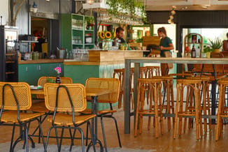 Modern café met gele rattan stoelen aan houten tafels, groene kasten, houten bar met barkrukken, hangende planten en losse we