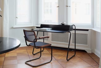 Zwart bureau met meshpaneel-voorkant aan raam, zwarte stoel met armleuningen en houten zitting, houten vloer, ronde tafel lin