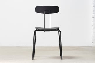 Zwarte stoel met houten zitting en rugleuning, metalen frame, staand tegen witte muur op betonnen vloer.