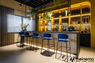 Modern bar met blauwe barkrukken, gele wandkasten met verlichting, groen, ramen met jaloeziën en betonnen vloer.