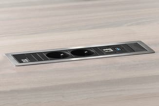 Zwart-grijs bureaublad power strip met twee stopcontacten, USB-poorten en schakelaar op licht houten tafelblad.