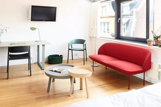 Kantoorruimte met rode sofa, twee ronde tafels, zwarte stoel, groene stoel, bureau met lamp en TV aan wand.