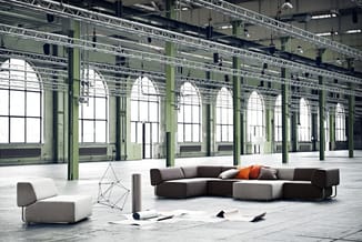 Modulaire grijze loungeset met oranje kussen in lege industriële hal met groene kolommen en grote ramen.