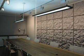 Vergaderruimte met lange houten tafel, zwarte stoelen, grijze 3D wandpanelen en zwarte hangende LED-verlichting.
