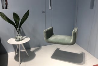 Showroom met grijs-groene hangstoel, wit bijzettafel met glazen vaas met groene planten en ME&U logo op wand.
