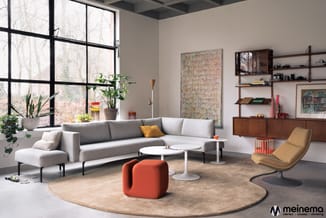 Modern kantoor/woonkamer met grijze hoekbank, gele fauteuil, rood poef, witte salontafels, houten kasten en grote ramen met u